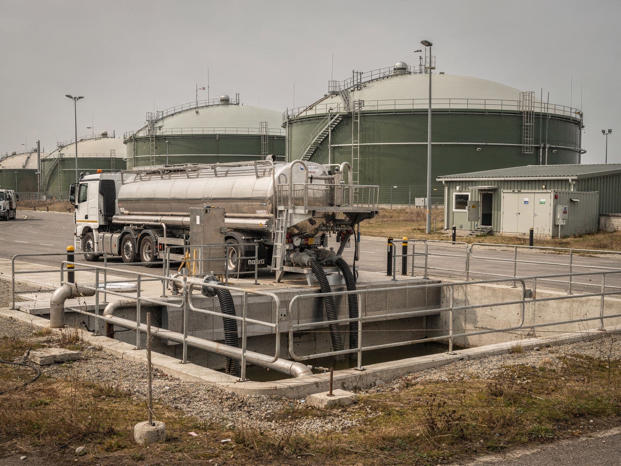 Tanker truck unloading feedstock at an anaerobic digestion facility, with digesters in the background.